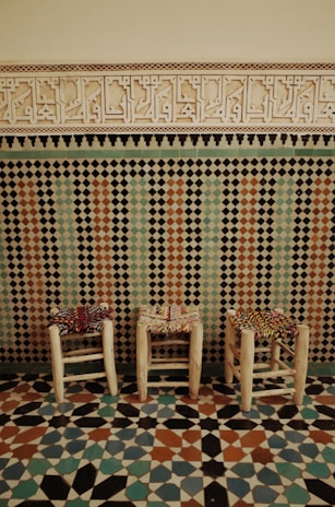 Three wooden stools with woven multicolored seats are positioned in front of an ornate wall. The wall features a geometric mosaic pattern, blending shades of blue, green, and rust, topped with intricate carved designs. The floor reflects similar vibrant patterns in a mosaic style.