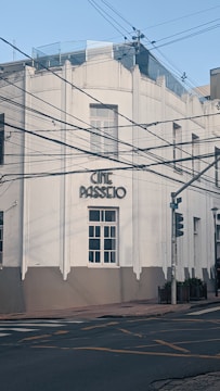 A white building with art deco architecture and the words 'Cine Passeio' displayed prominently. The facade features large windows, and the building is situated at a street corner with traffic lights. Overhead power lines crisscross the scene, adding an urban element.