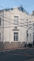 A white building with art deco architecture and the words 'Cine Passeio' displayed prominently. The facade features large windows, and the building is situated at a street corner with traffic lights. Overhead power lines crisscross the scene, adding an urban element.