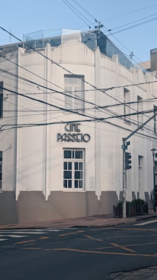 A white building with art deco architecture and the words 'Cine Passeio' displayed prominently. The facade features large windows, and the building is situated at a street corner with traffic lights. Overhead power lines crisscross the scene, adding an urban element.