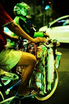 A person on a bicycle is selling various colorful packaged goods hanging from the handlebars. Another person wearing a green helmet and jacket is riding a motorcycle in the background. The scene takes place on a street at night, creating a vibrant and bustling atmosphere.