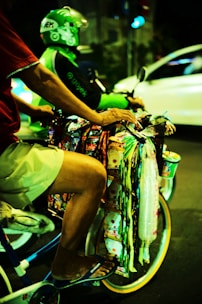 A person on a bicycle is selling various colorful packaged goods hanging from the handlebars. Another person wearing a green helmet and jacket is riding a motorcycle in the background. The scene takes place on a street at night, creating a vibrant and bustling atmosphere.