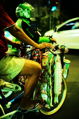 A person on a bicycle is selling various colorful packaged goods hanging from the handlebars. Another person wearing a green helmet and jacket is riding a motorcycle in the background. The scene takes place on a street at night, creating a vibrant and bustling atmosphere.