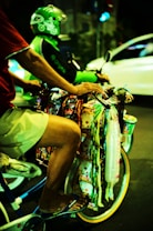 A person on a bicycle is selling various colorful packaged goods hanging from the handlebars. Another person wearing a green helmet and jacket is riding a motorcycle in the background. The scene takes place on a street at night, creating a vibrant and bustling atmosphere.