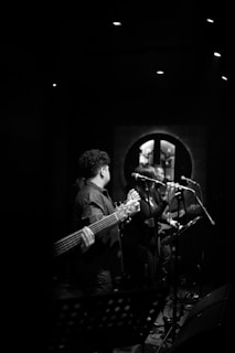 Musicians performing on stage in a dimly lit venue, with a focus on a guitarist holding a bass guitar and a vocalist singing into a microphone. The background includes soft lighting and a decorative wall element, creating a moody atmosphere.