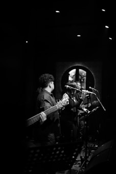 Musicians performing on stage in a dimly lit venue, with a focus on a guitarist holding a bass guitar and a vocalist singing into a microphone. The background includes soft lighting and a decorative wall element, creating a moody atmosphere.