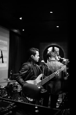 A black and white photograph of a musician holding a guitar on stage. The setting appears to be a dimly lit live performance venue with a drum set visible in the background. The musician is focused on their performance, and there is a microphone stand in front of them.
