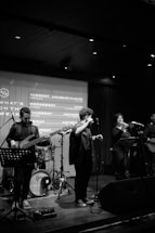 A live music performance with multiple musicians on stage. The scene includes a bassist, a vocalist with a microphone, and additional performers, each deeply engaged in the music. The setting is dimly lit, with spotlights illuminating the stage. An event schedule is projected onto a screen in the background.