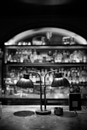 A dimly lit bar counter featuring a vintage-style double lamp with two curved metal shades. The background displays a blurred view of shelves stocked with various bottles. There is a sign with a QR code next to the lamp on the counter.
