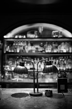 A dimly lit bar counter featuring a vintage-style double lamp with two curved metal shades. The background displays a blurred view of shelves stocked with various bottles. There is a sign with a QR code next to the lamp on the counter.