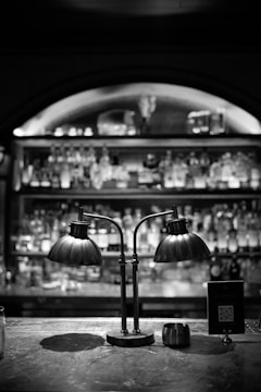 A dimly lit bar counter featuring a vintage-style double lamp with two curved metal shades. The background displays a blurred view of shelves stocked with various bottles. There is a sign with a QR code next to the lamp on the counter.