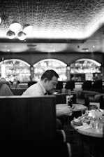 A person is seated at a table in a restaurant or café, focusing intently on their smartphone. The scene includes multiple tables with glassware, a modern design with a textured ceiling, decorative lighting, and a bar in the background with shelves displaying various bottles.