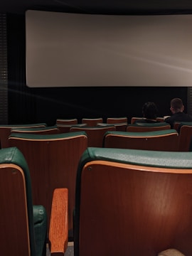 A dimly lit movie theater with green and wooden seats. The theater screen is blank, and only a few people are sitting towards the front, creating a secluded and quiet atmosphere.