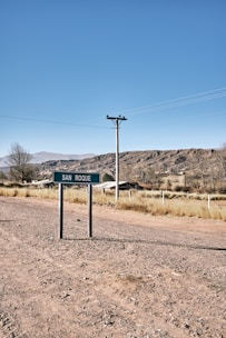 A rural landscape with a road sign labeled 'San Roque' in the foreground. The scene includes a dirt road, some grasses, a utility pole with wires, and a house in the background. The environment is open, with dry ground and distant hills under a clear blue sky.