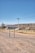 A rural landscape with a road sign labeled 'San Roque' in the foreground. The scene includes a dirt road, some grasses, a utility pole with wires, and a house in the background. The environment is open, with dry ground and distant hills under a clear blue sky.