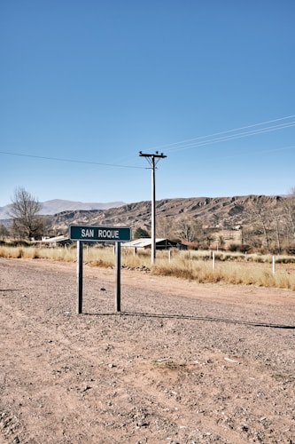A rural landscape with a road sign labeled 'San Roque' in the foreground. The scene includes a dirt road, some grasses, a utility pole with wires, and a house in the background. The environment is open, with dry ground and distant hills under a clear blue sky.
