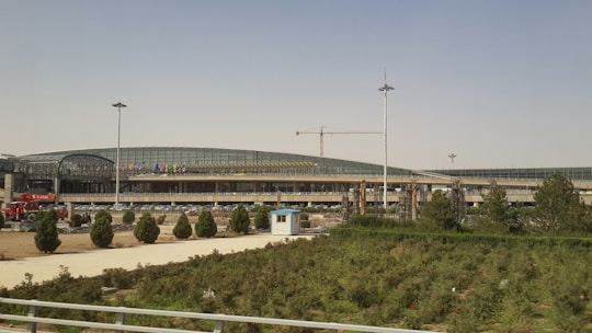 A modern architectural structure with a curved roof and extensive glass panels. Several trees and a patch of grass are in the foreground. There is a crane visible, indicating ongoing construction work.