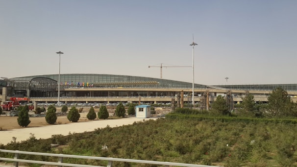 A modern architectural structure with a curved roof and extensive glass panels. Several trees and a patch of grass are in the foreground. There is a crane visible, indicating ongoing construction work.