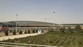 A modern architectural structure with a curved roof and extensive glass panels. Several trees and a patch of grass are in the foreground. There is a crane visible, indicating ongoing construction work.