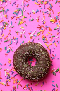 A chocolate frosted donut with colorful sprinkles is placed on a pink background filled with scattered multicolored sprinkles.