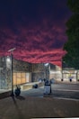 A modern building under a dramatic, colorful sky with vibrant pink and purple clouds. The structure features large windows and artistic stonework, with a lit pathway and outdoor lamps. A sign indicates the direction to a university store. Potted plants adorn the entrance.