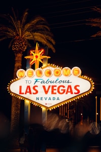A brightly illuminated sign welcomes visitors to Las Vegas. The sign features classic retro styling with the words 'Welcome to Fabulous Las Vegas Nevada' highlighted in vibrant lights. Palm trees can be seen in the background, with the scene set during nighttime, creating a lively and inviting atmosphere.