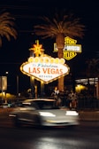 A bright and iconic 'Welcome to Fabulous Las Vegas, Nevada' sign is illuminated at night, surrounded by palm trees. A blurred white car passes by in the foreground and a group of people gathers nearby. Neon lights and a Harley-Davidson sign are visible in the background.