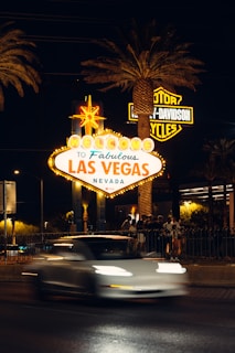 A bright and iconic 'Welcome to Fabulous Las Vegas, Nevada' sign is illuminated at night, surrounded by palm trees. A blurred white car passes by in the foreground and a group of people gathers nearby. Neon lights and a Harley-Davidson sign are visible in the background.