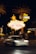 A bright and iconic 'Welcome to Fabulous Las Vegas, Nevada' sign is illuminated at night, surrounded by palm trees. A blurred white car passes by in the foreground and a group of people gathers nearby. Neon lights and a Harley-Davidson sign are visible in the background.
