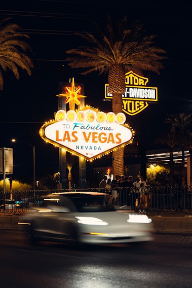 A bright and iconic 'Welcome to Fabulous Las Vegas, Nevada' sign is illuminated at night, surrounded by palm trees. A blurred white car passes by in the foreground and a group of people gathers nearby. Neon lights and a Harley-Davidson sign are visible in the background.