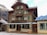 A traditional wooden chalet-style building with green shutters and a large, covered patio. The architecture is rustic with intricate woodwork. Snow-dusted mountains are visible in the background, suggesting a cold, alpine setting. Two white benches are placed on the patio.