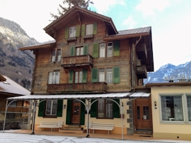 A traditional wooden chalet-style building with green shutters and a large, covered patio. The architecture is rustic with intricate woodwork. Snow-dusted mountains are visible in the background, suggesting a cold, alpine setting. Two white benches are placed on the patio.