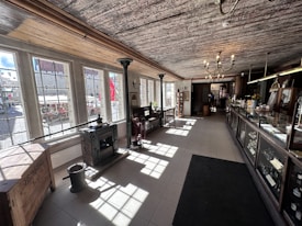 A vintage interior with large windows allowing natural light to fill the space. The room features dark wooden furnishings, including ornate chandeliers, antique machinery, and display cabinets along one side. The floor is tiled, and there are various decorative objects throughout the room.