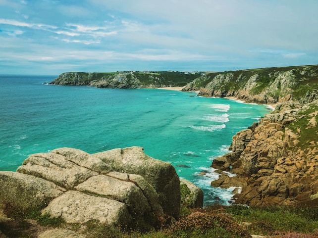 A scenic coastal landscape featuring turquoise waters gently lapping against rugged cliffs. The coastline stretches into the distance under a partly cloudy sky. The foreground includes rocky terrain with patches of green vegetation.