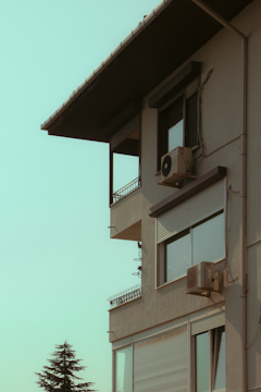 A modern building with large windows and air conditioning units mounted on the outer walls, set against a clear sky. A tree is visible in the foreground, adding a natural element to the urban scene.