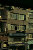 A multi-story building with various business signage, including one that reads 'Taksim Büro Kırtasiye.' The facade shows multiple windows and a few air conditioning units attached to the exterior. The building's exterior appears slightly worn, and some balconies and old metal railings are visible. The overall scene is dimly lit, probably captured during early morning or late afternoon.