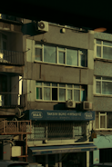A multi-story building with various business signage, including one that reads 'Taksim Büro Kırtasiye.' The facade shows multiple windows and a few air conditioning units attached to the exterior. The building's exterior appears slightly worn, and some balconies and old metal railings are visible. The overall scene is dimly lit, probably captured during early morning or late afternoon.