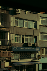 A multi-story building with various business signage, including one that reads 'Taksim Büro Kırtasiye.' The facade shows multiple windows and a few air conditioning units attached to the exterior. The building's exterior appears slightly worn, and some balconies and old metal railings are visible. The overall scene is dimly lit, probably captured during early morning or late afternoon.