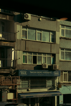 A multi-story building with various business signage, including one that reads 'Taksim Büro Kırtasiye.' The facade shows multiple windows and a few air conditioning units attached to the exterior. The building's exterior appears slightly worn, and some balconies and old metal railings are visible. The overall scene is dimly lit, probably captured during early morning or late afternoon.