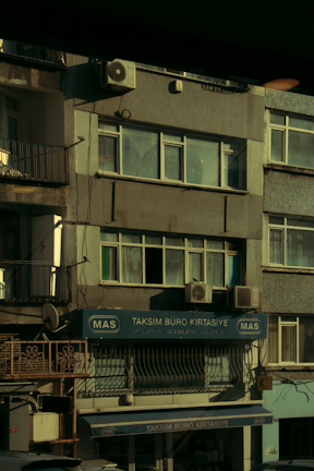 A multi-story building with various business signage, including one that reads 'Taksim Büro Kırtasiye.' The facade shows multiple windows and a few air conditioning units attached to the exterior. The building's exterior appears slightly worn, and some balconies and old metal railings are visible. The overall scene is dimly lit, probably captured during early morning or late afternoon.