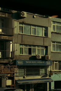A multi-story building with various business signage, including one that reads 'Taksim Büro Kırtasiye.' The facade shows multiple windows and a few air conditioning units attached to the exterior. The building's exterior appears slightly worn, and some balconies and old metal railings are visible. The overall scene is dimly lit, probably captured during early morning or late afternoon.