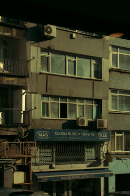 A multi-story building with various business signage, including one that reads 'Taksim B&uuml;ro Kırtasiye.' The facade shows multiple windows and a few air conditioning units attached to the exterior. The building's exterior appears slightly worn, and some balconies and old metal railings are visible. The overall scene is dimly lit, probably captured during early morning or late afternoon.