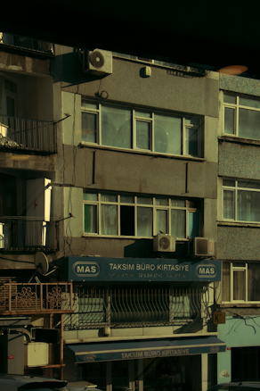 A multi-story building with various business signage, including one that reads 'Taksim B&uuml;ro Kırtasiye.' The facade shows multiple windows and a few air conditioning units attached to the exterior. The building's exterior appears slightly worn, and some balconies and old metal railings are visible. The overall scene is dimly lit, probably captured during early morning or late afternoon.