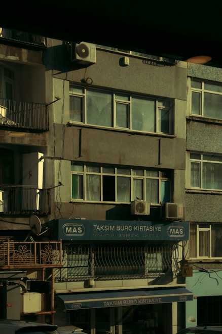 A multi-story building with various business signage, including one that reads 'Taksim B&uuml;ro Kırtasiye.' The facade shows multiple windows and a few air conditioning units attached to the exterior. The building's exterior appears slightly worn, and some balconies and old metal railings are visible. The overall scene is dimly lit, probably captured during early morning or late afternoon.