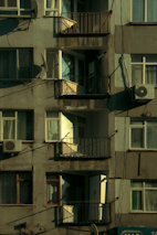 A multi-story residential building facade featuring balconies, windows, and satellite dishes. The paint appears weathered, and there are visible air conditioning units attached to some windows. Sunlight casts shadows across the surface, highlighting the textures of the walls and railings.
