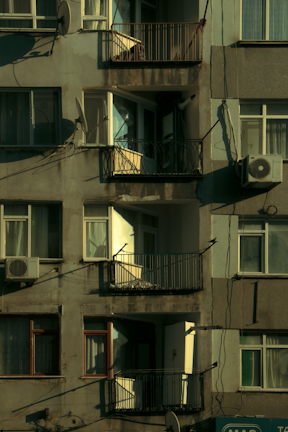A multi-story residential building facade featuring balconies, windows, and satellite dishes. The paint appears weathered, and there are visible air conditioning units attached to some windows. Sunlight casts shadows across the surface, highlighting the textures of the walls and railings.
