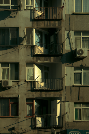 A multi-story residential building facade featuring balconies, windows, and satellite dishes. The paint appears weathered, and there are visible air conditioning units attached to some windows. Sunlight casts shadows across the surface, highlighting the textures of the walls and railings.