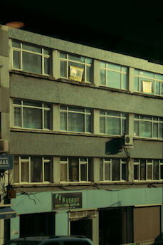A multi-story building with a retro design features numerous rectangular windows. An air conditioning unit is affixed to the exterior wall, and several signs and advertisements are displayed on the ground floor. The structure appears to have a slightly weathered, urban appearance.
