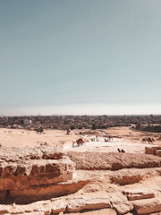 A vast desert landscape with sandy terrain stretching into the distance. In the foreground, large stone blocks hint at ancient ruins. Several camels and people are scattered across the area, with a bustling city skyline visible in the background under a clear blue sky.