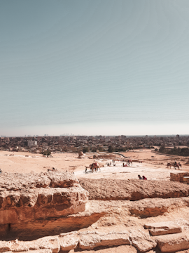 A vast desert landscape with sandy terrain stretching into the distance. In the foreground, large stone blocks hint at ancient ruins. Several camels and people are scattered across the area, with a bustling city skyline visible in the background under a clear blue sky.